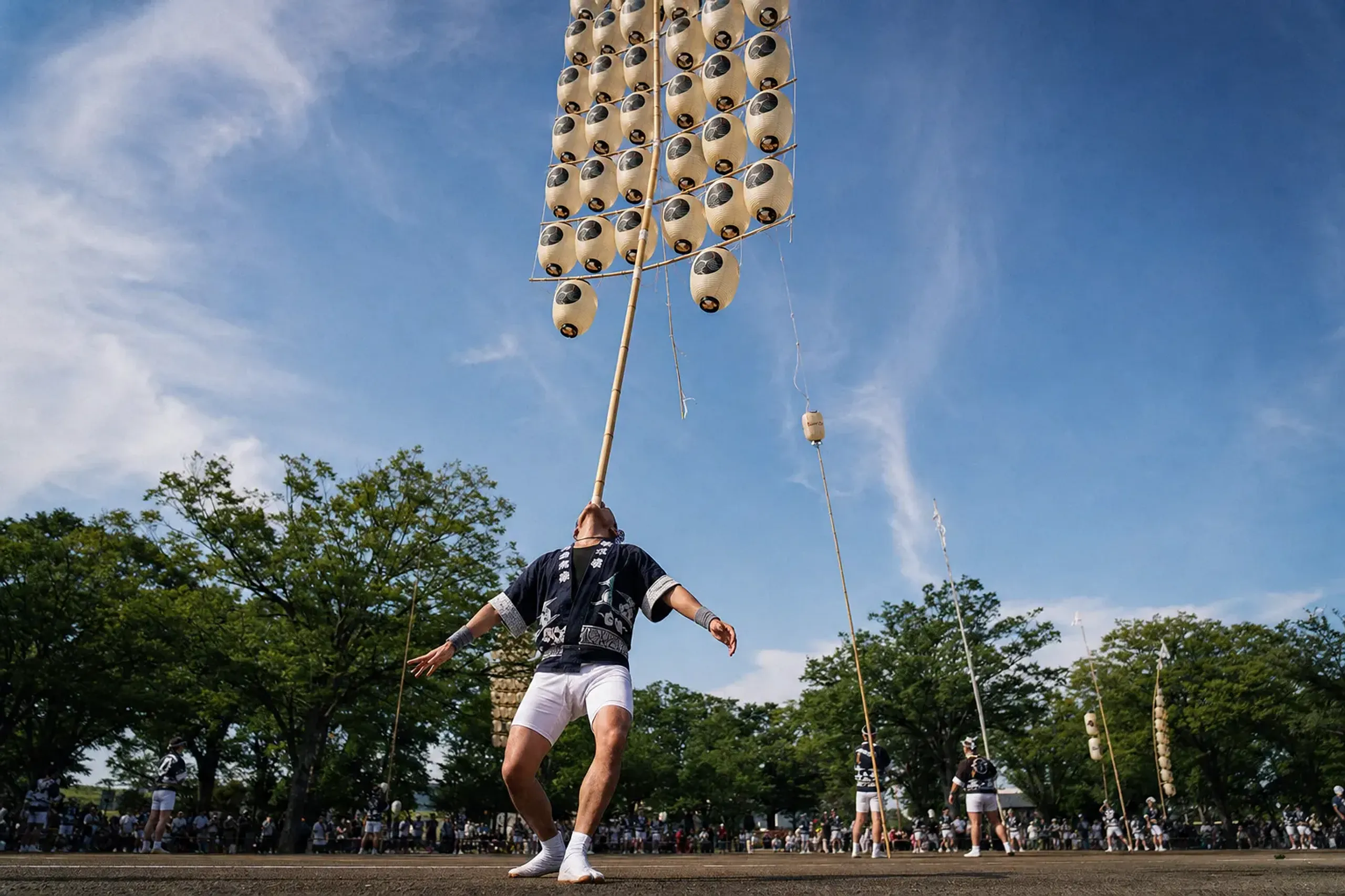 A single sashite at the daytime Kantō Myōgi Taikai balancing a 12-meter bamboo Kantō pole on his forehead in bright sunlight, feet mid-adjustment