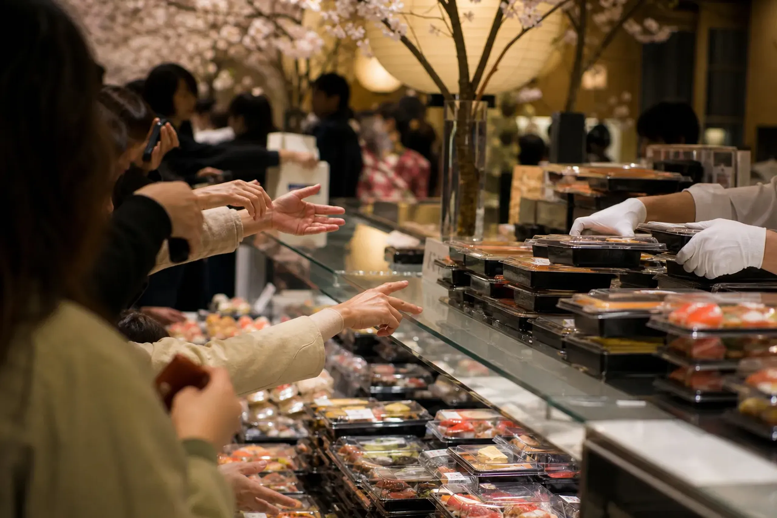 A prepared-food counter in the evening with multiple takeaway trays being restocked while a dense cluster of shoppers’ hands reach in—capturing the “markdown hunt” atmosphere through crowded proximity