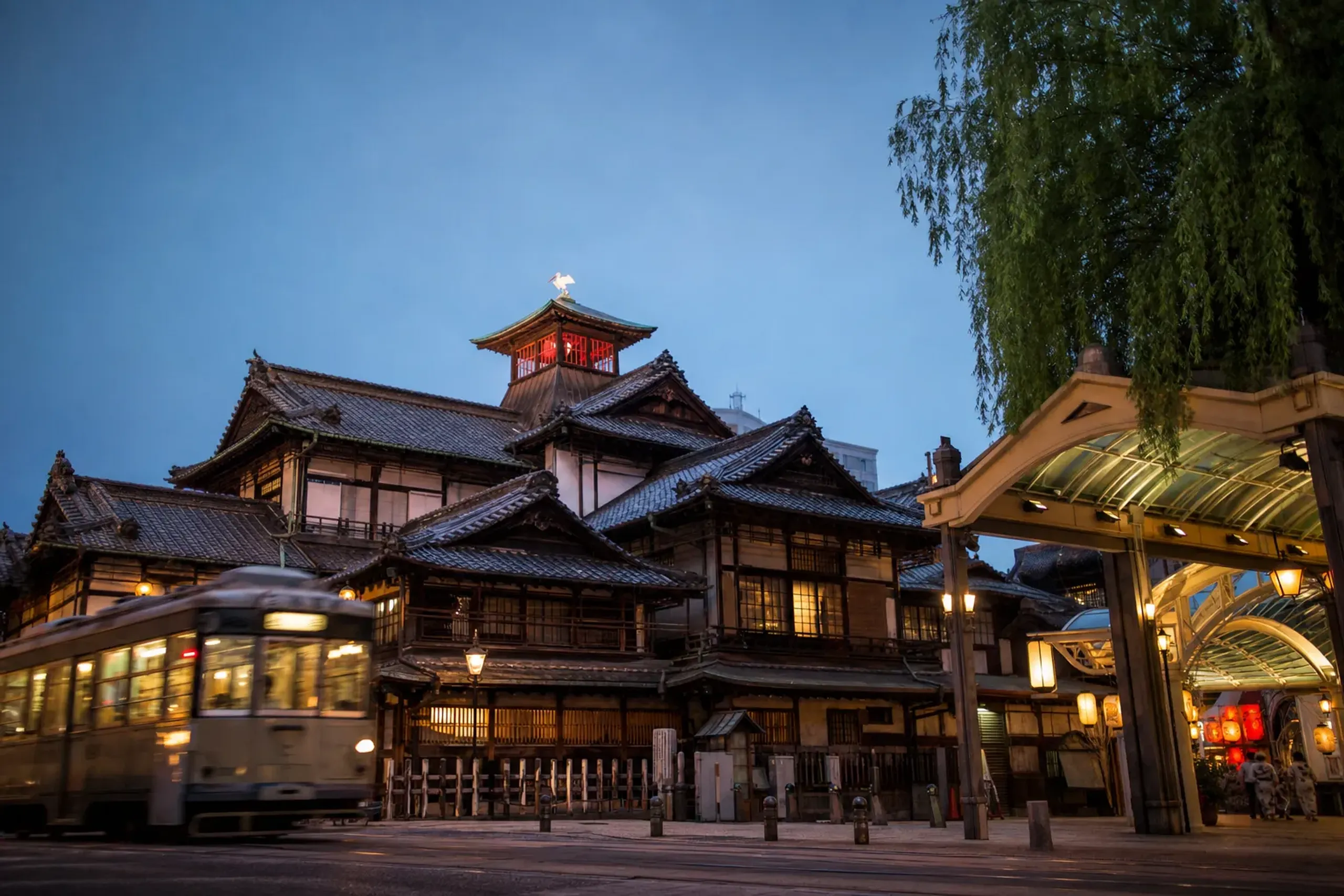 the historic wooden Dōgo Onsen bathhouse at the centre of Matsuyama, with a passing tram and the entrance to the covered shopping arcade nearby at dusk