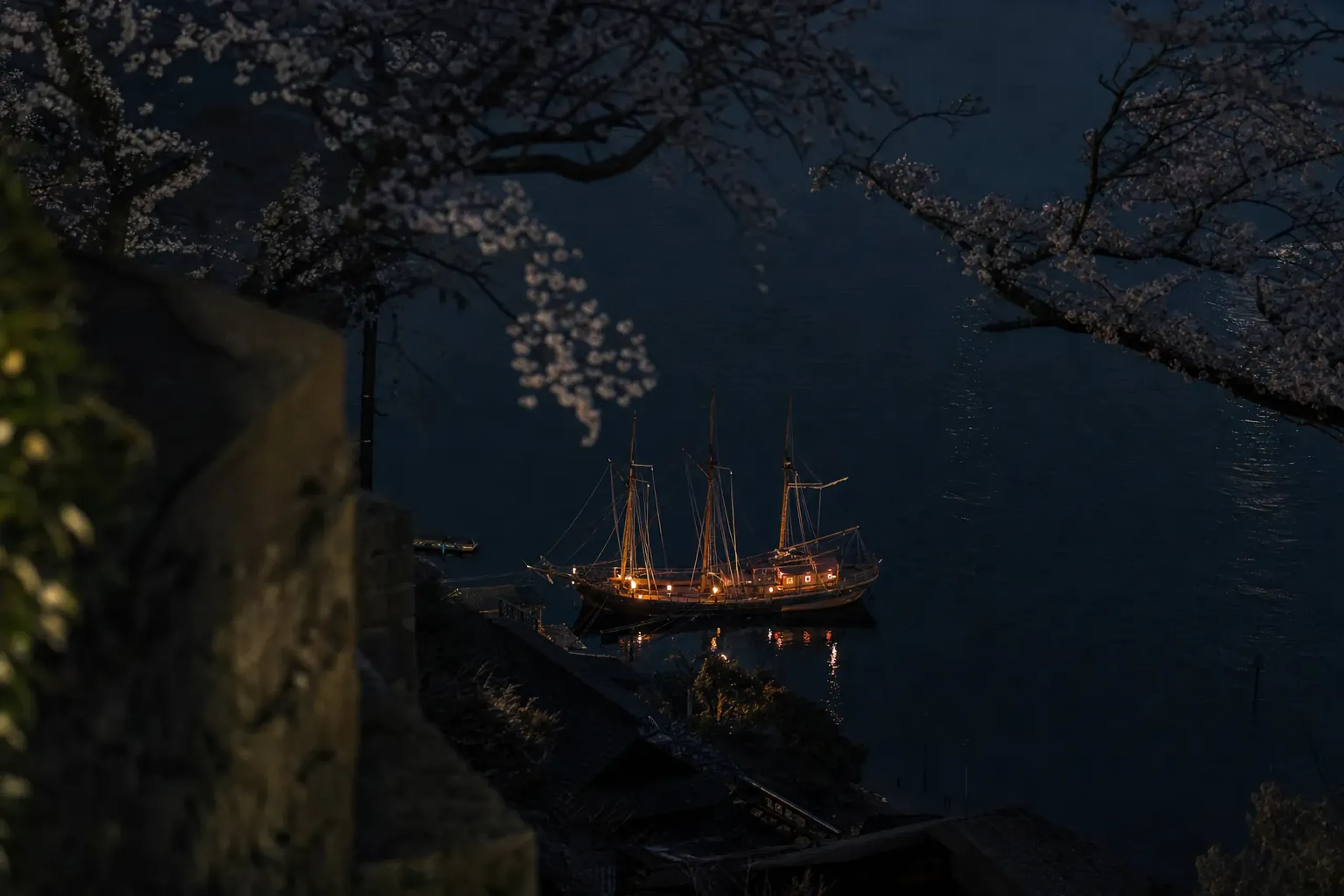 A wooden sailing ship at anchor in Nagasaki harbor at night, seen from the Minami-Yamate hillside, lantern light reflecting on dark water