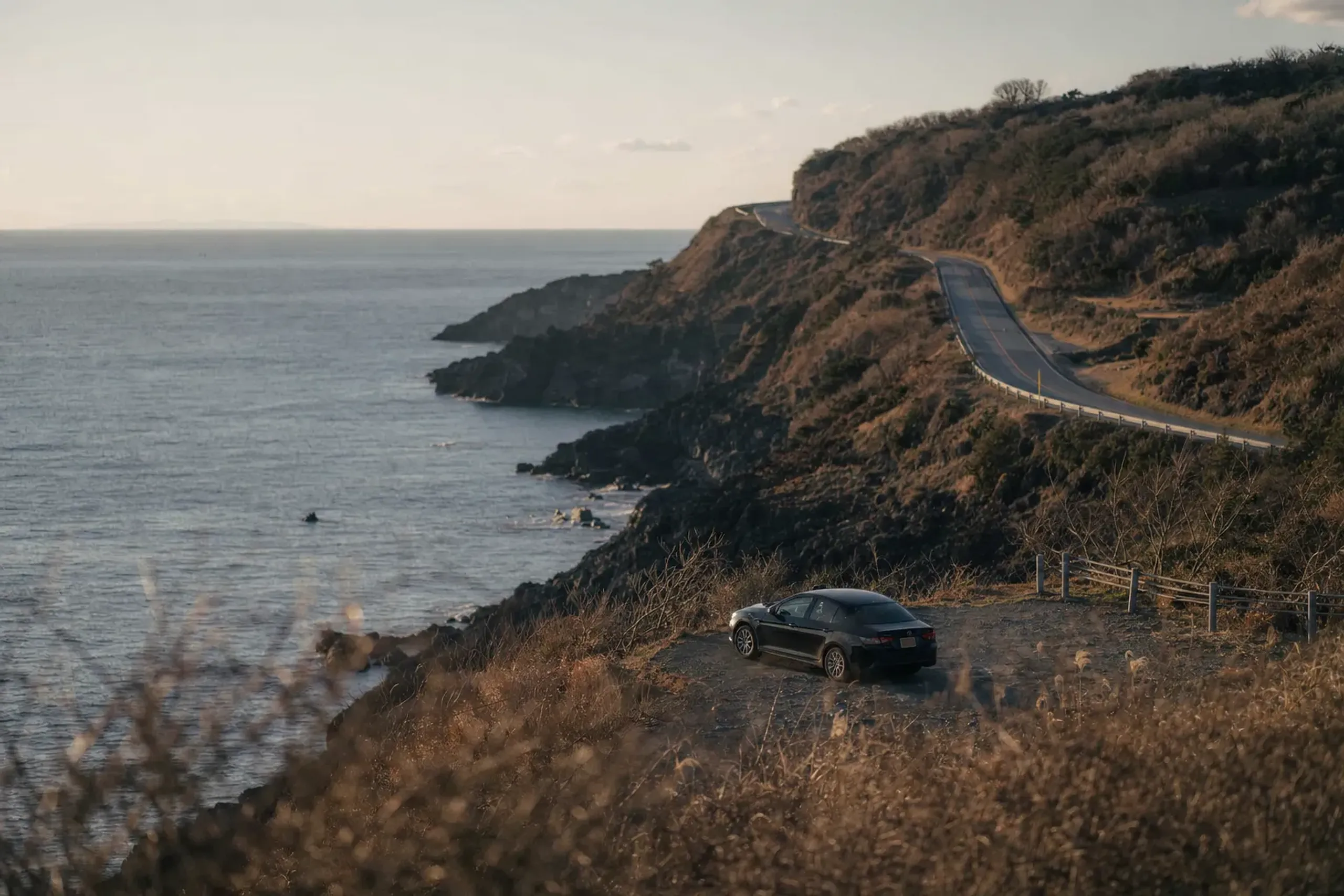 The western coast road of Fukue Island winding along rugged East China Sea cliffs at golden hour, with a lone private vehicle parked at a scenic viewpoint