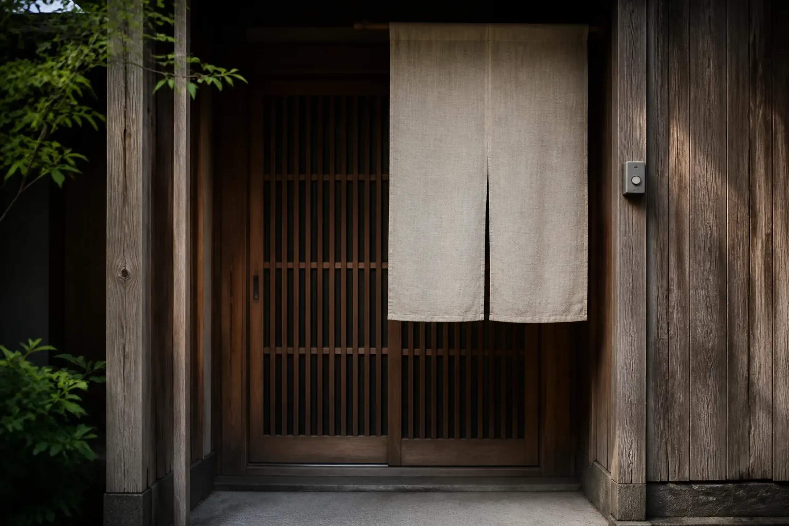 Exterior detail of a discreet craft studio entrance in Japan: a plain noren curtain at the doorway, an unmarked sliding wooden door, and a small, unlabeled wall-mounted doorbell beside it—no signage v
