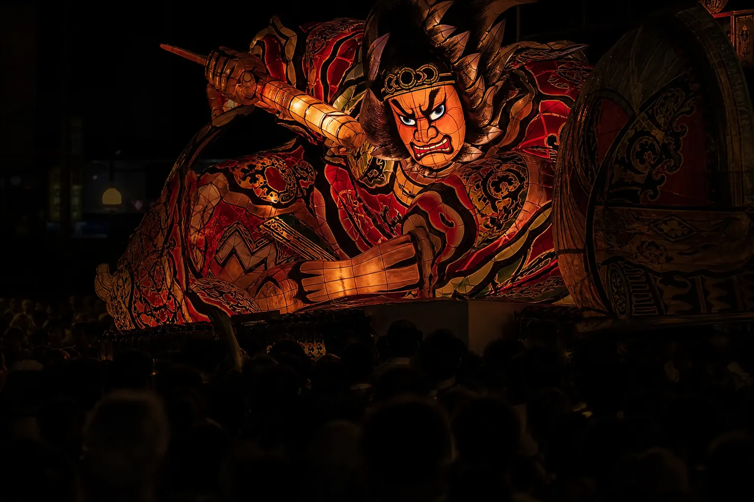 an illuminated Nebuta float glowing against dark Aomori streets as it turns with engineered weight through a shoulder-to-shoulder evening crowd