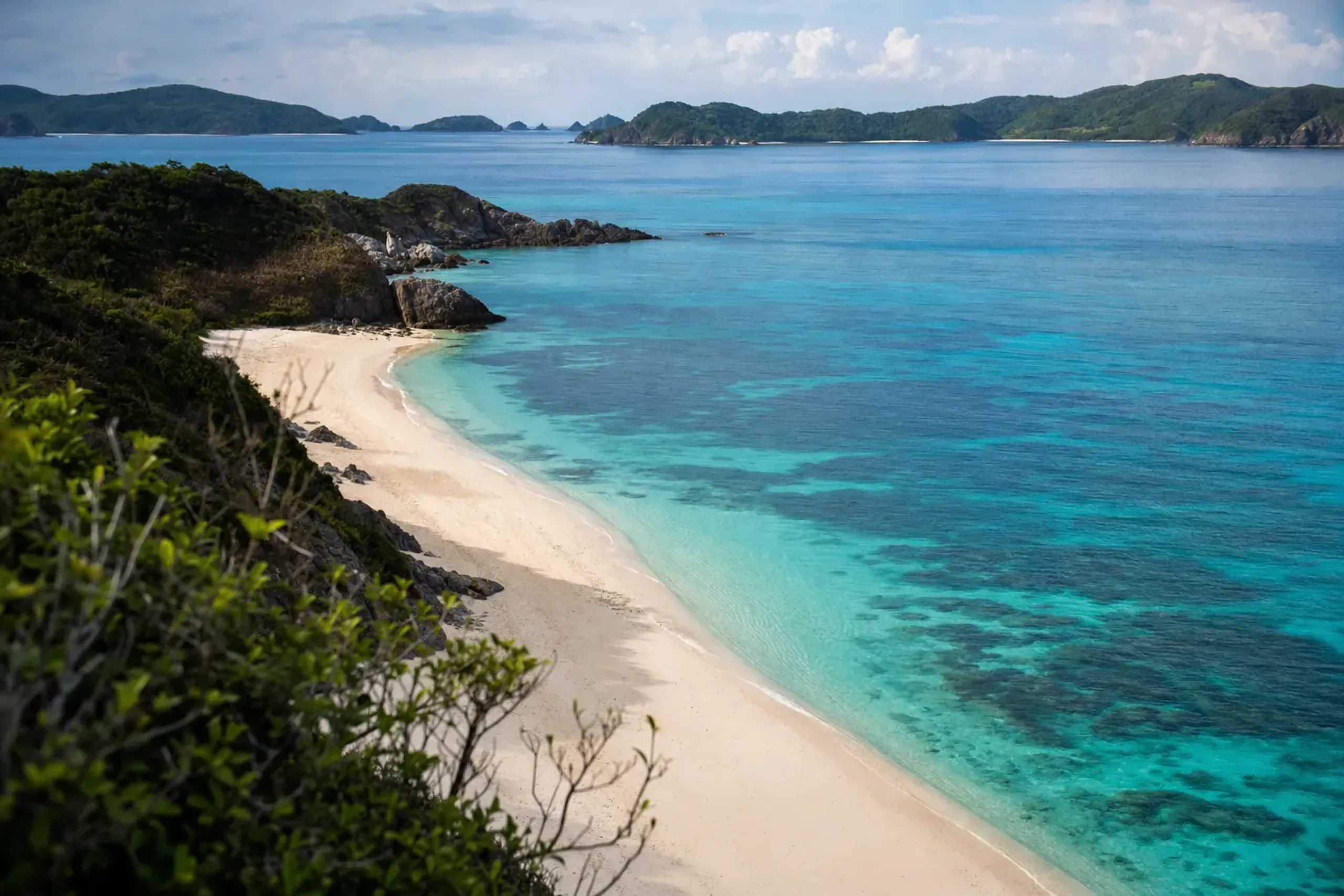 the broad pale-sand curve of Furuzamami Beach on Zamami Island with its reef visible just offshore in turquoise water