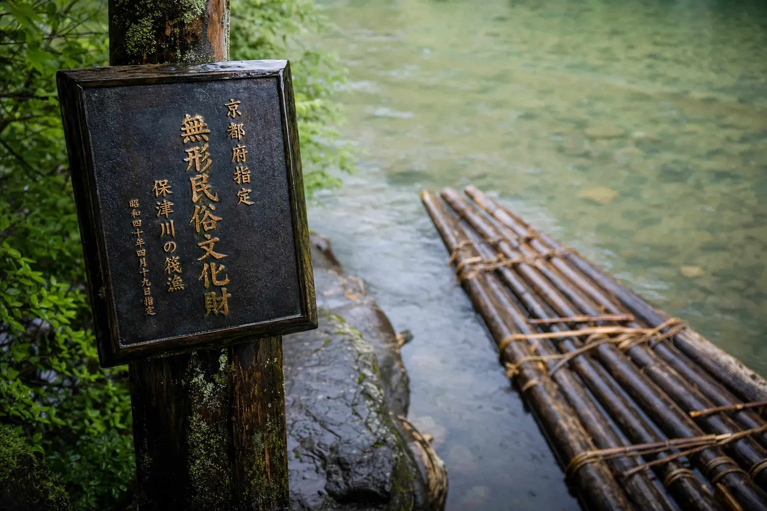 Kitayama Prefecture's official certificate plaque designating log-raft building as an Intangible Folk Cultural Property, mounted beside a cedar raft on the Kitayama River