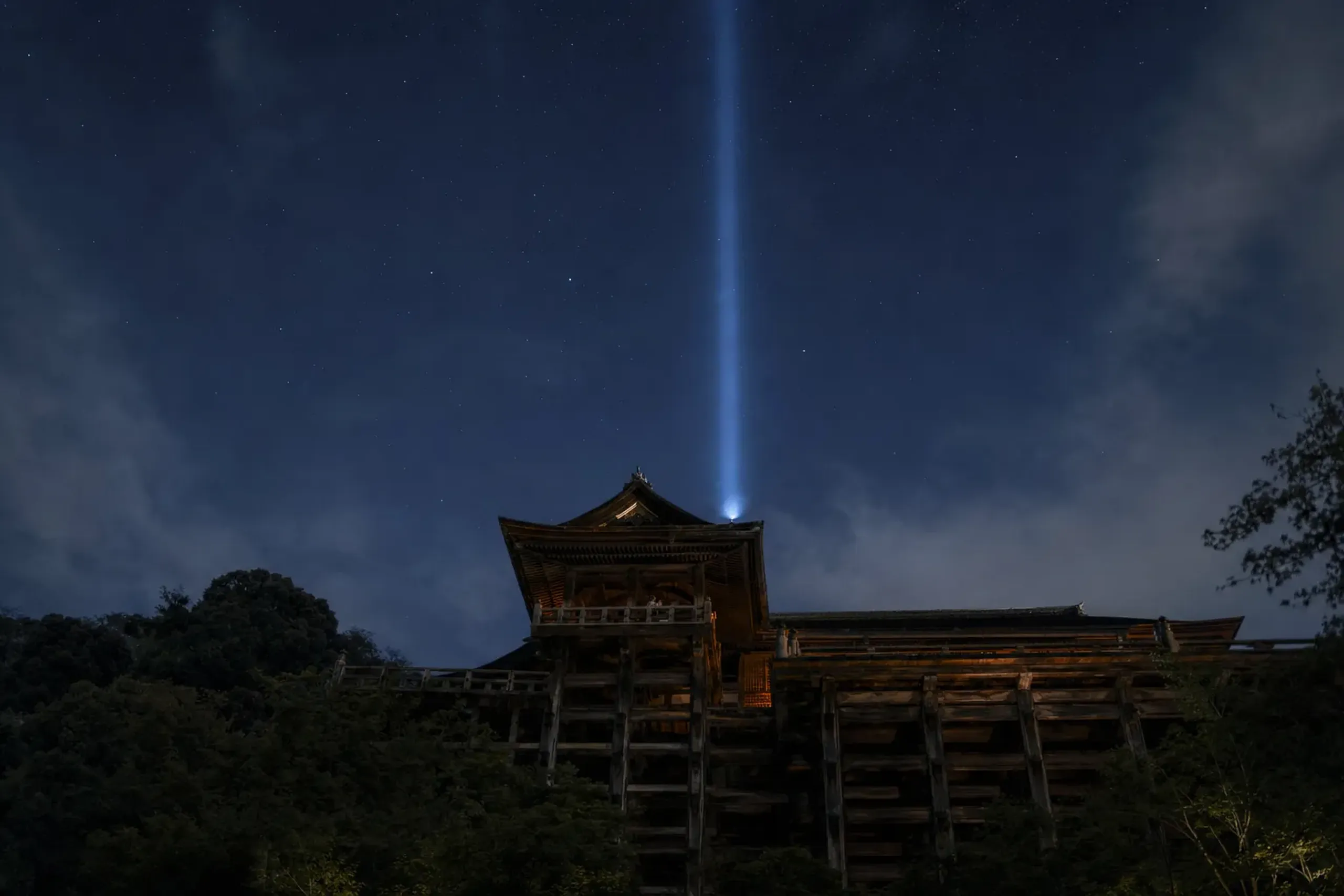 The blue column of Kannon Jihi-kō light rising from the darkened wooden hall of Kiyomizu-dera into the night sky over Higashiyama