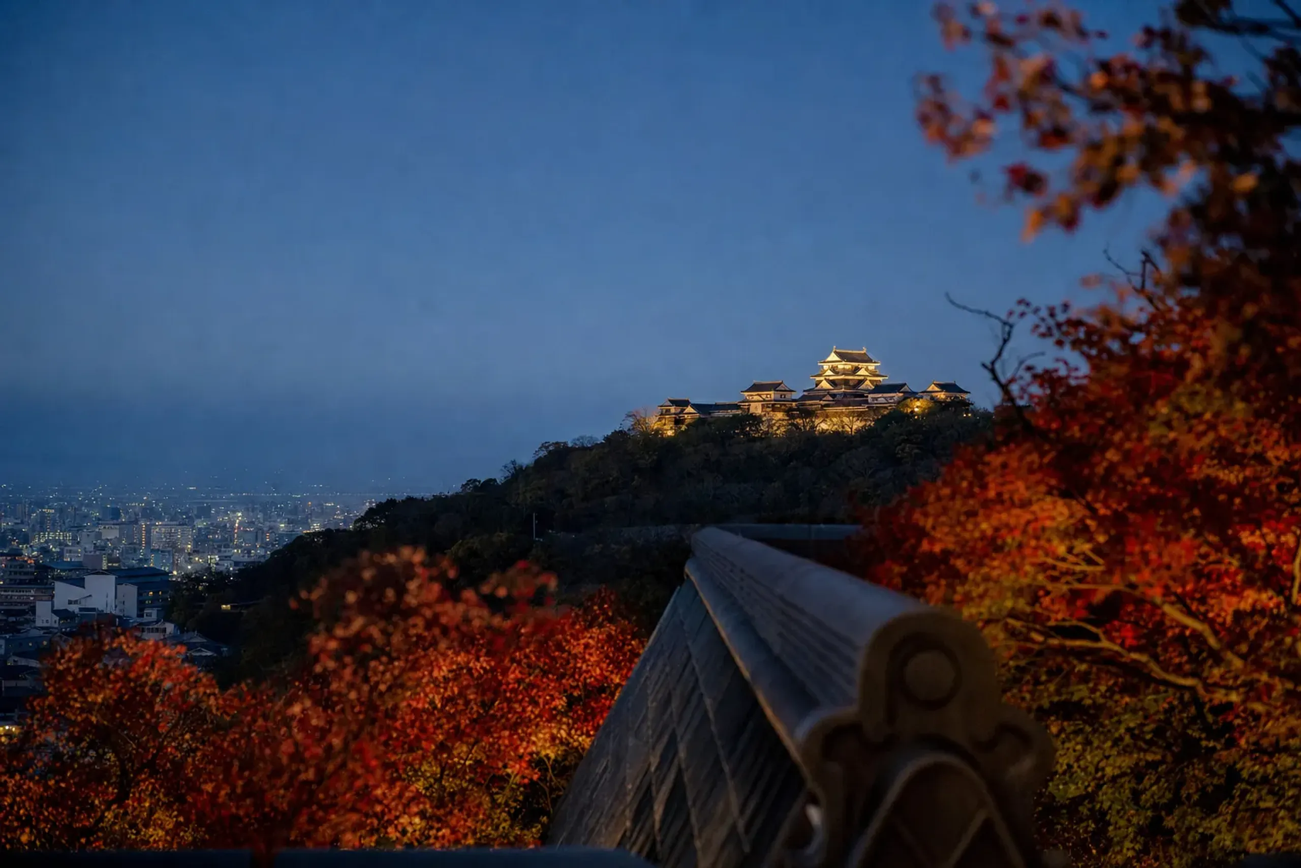 The keep of Matsuyama Castle illuminated at dusk above the darkened city grid of Matsuyama