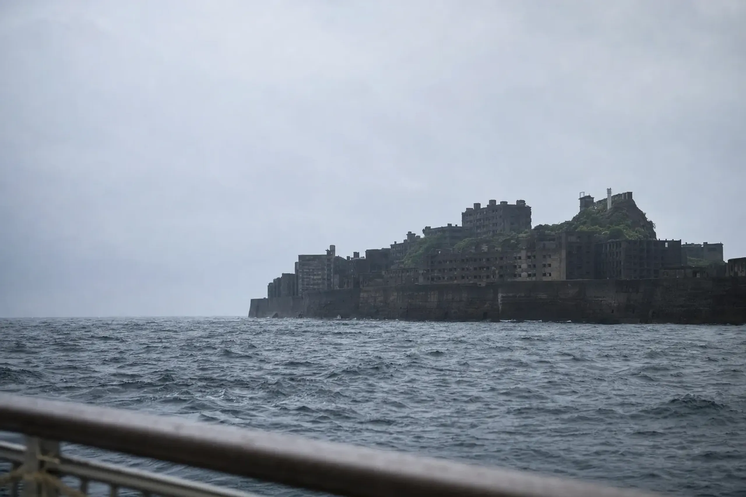 The silhouette of Hashima Island's ruined battleship-shaped skyline viewed across choppy harbor water from a Nagasaki cruise boat on an overcast day