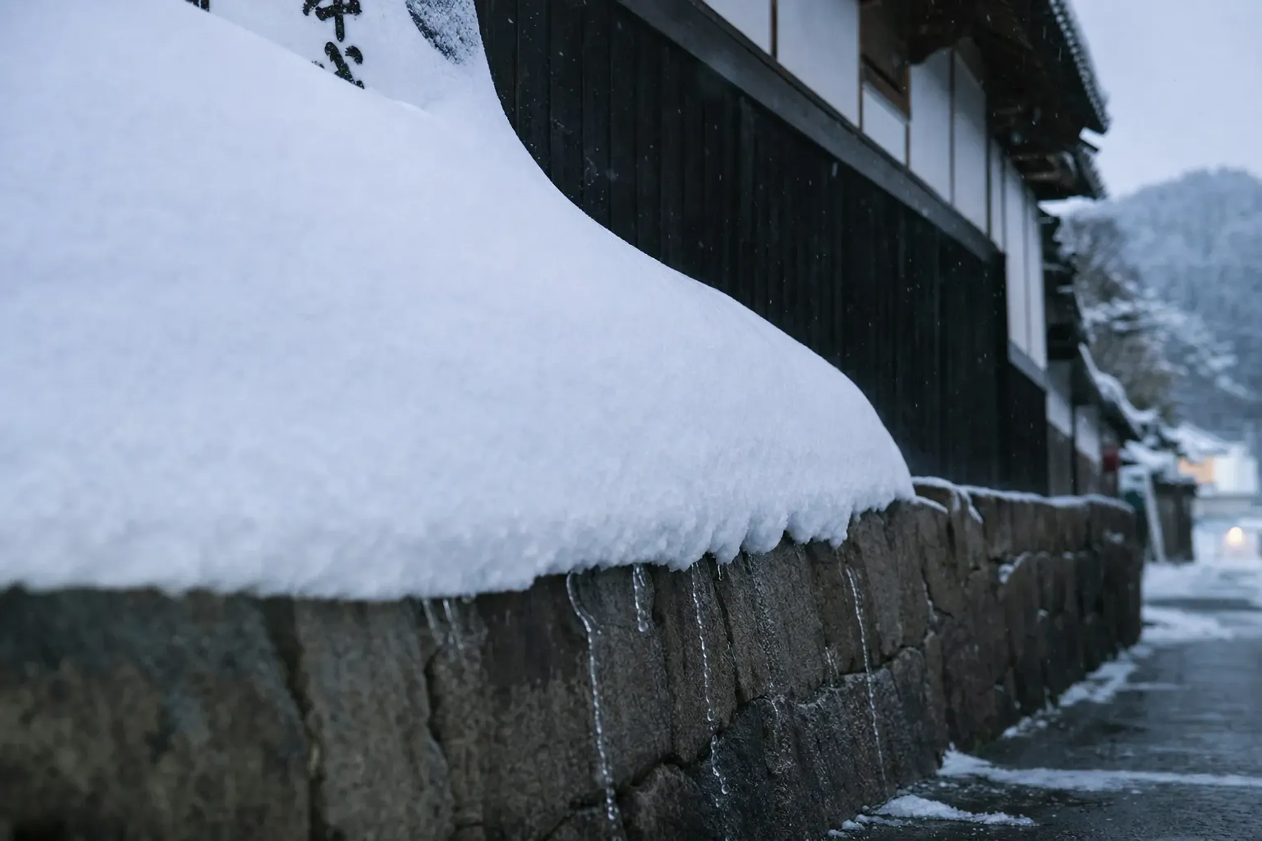Deep winter snowdrifts piled against a Niigata brewery wall, with meltwater seeping through weathered granite rock