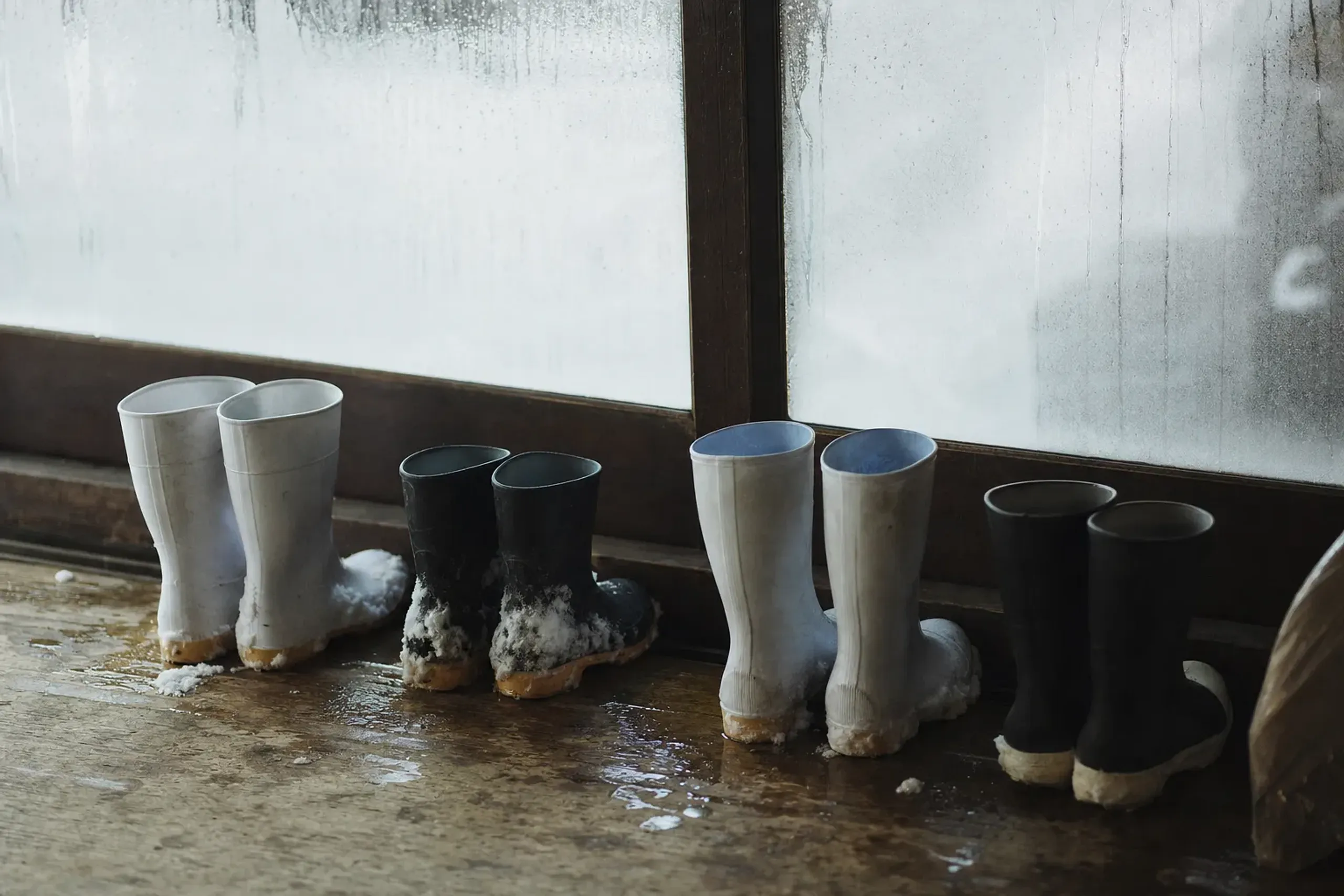 Wet boots lined up at a brewery threshold beside steam-fogged windows in a working winter sake brewery