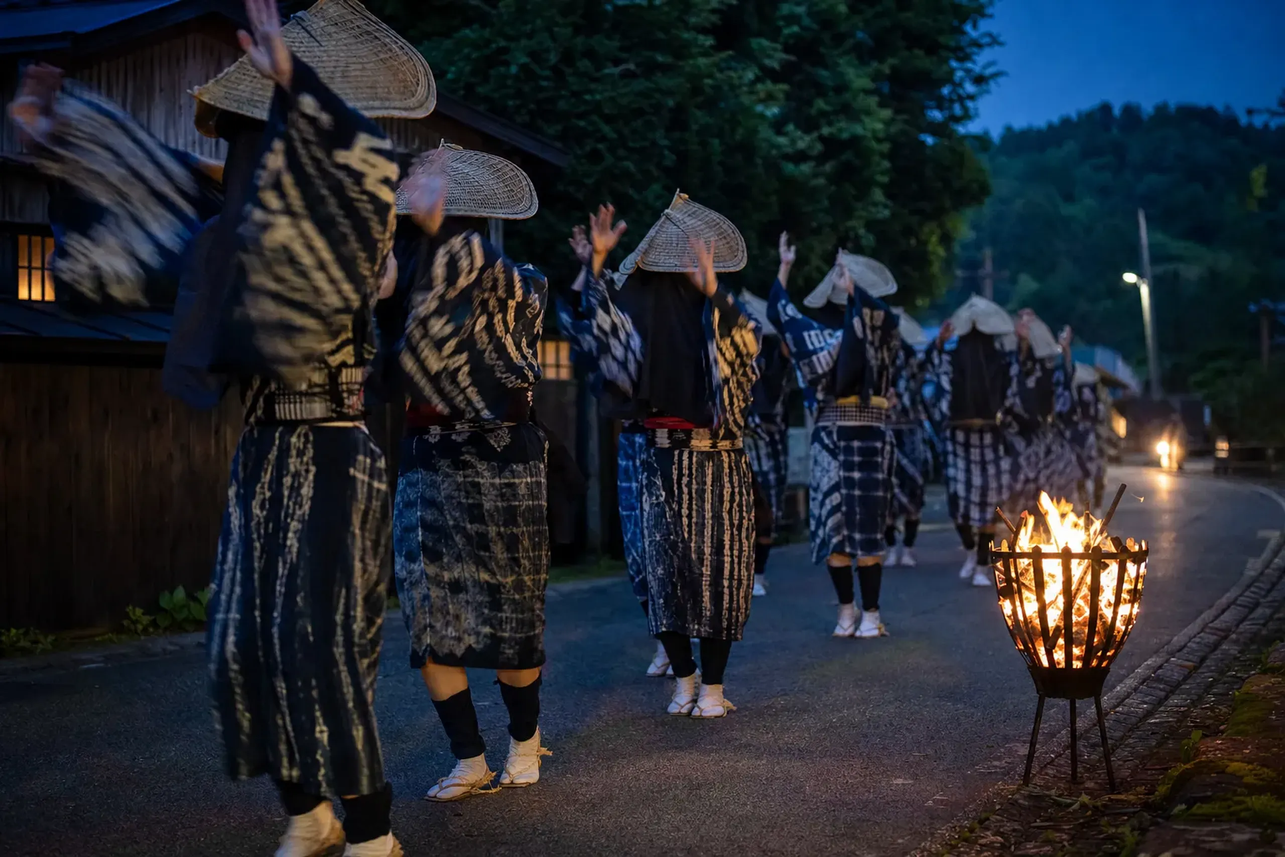 Hooded dancers in indigo shibori-dyed yukata gliding in a single file loop around the bonfires along Nishimonai Honcho Street, arms rising in liquid motion