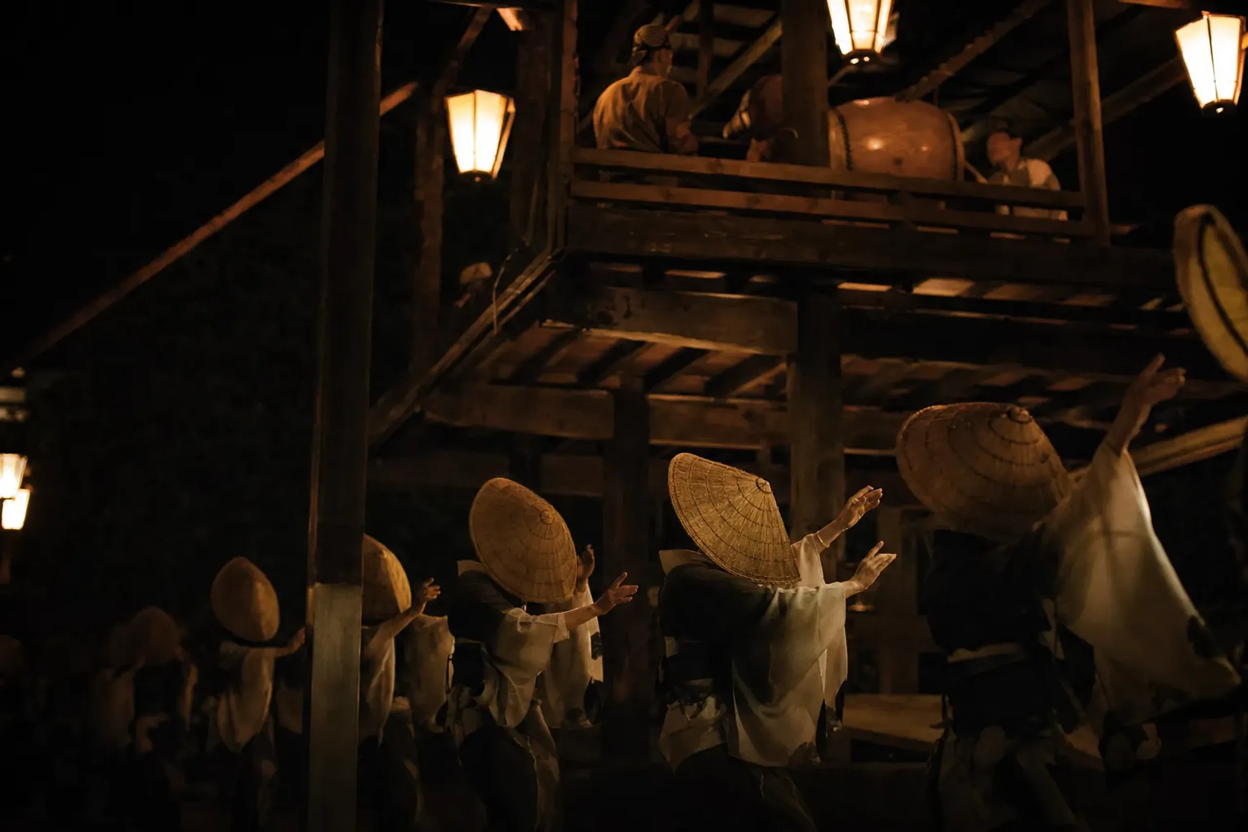 Adult women dancers in deep woven hats tilted low, dancing beneath the raised wooden musicians' scaffold at night