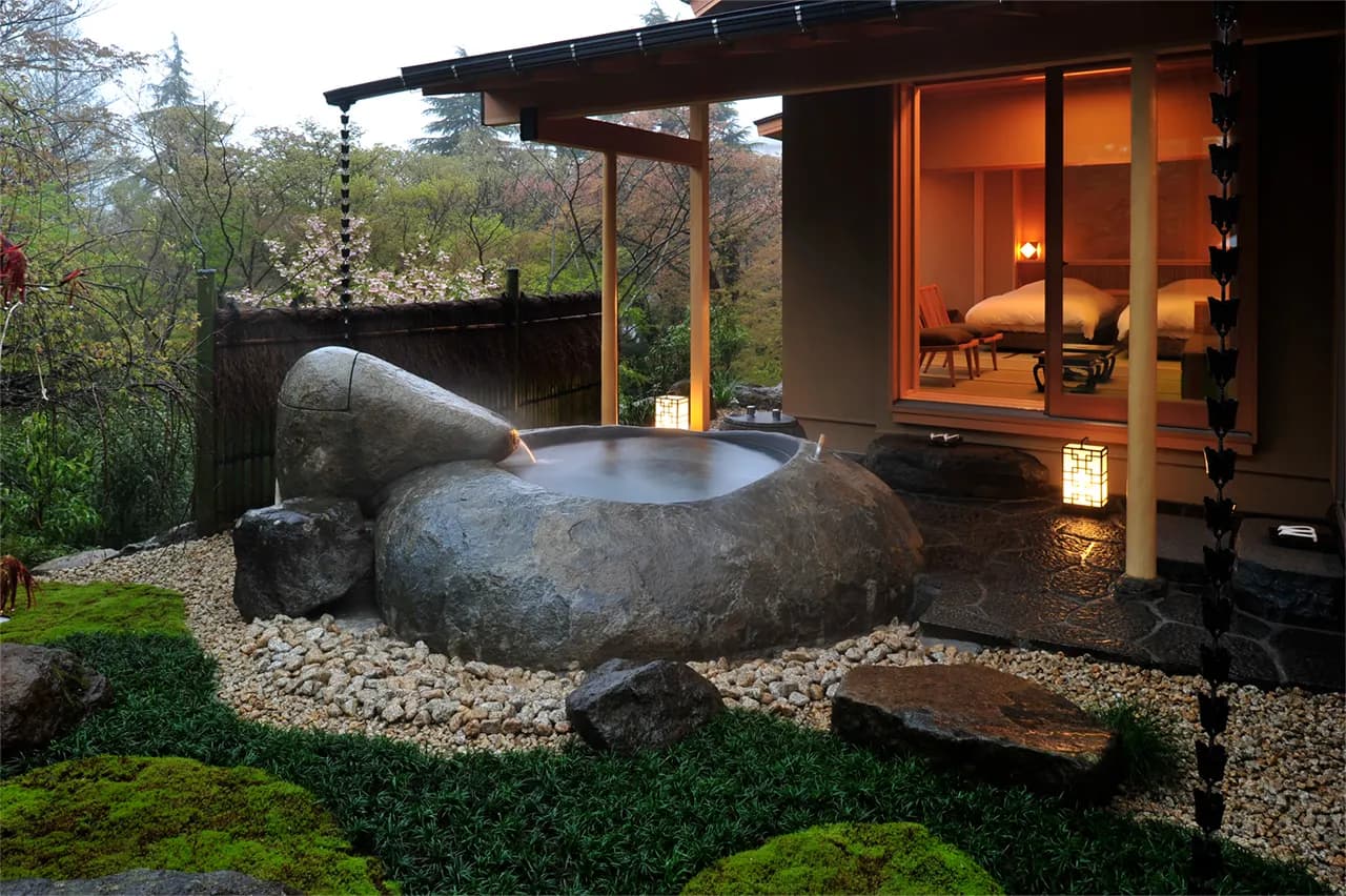 A private open-air onsen carved from rock, the lit ryokan suite glowing behind it