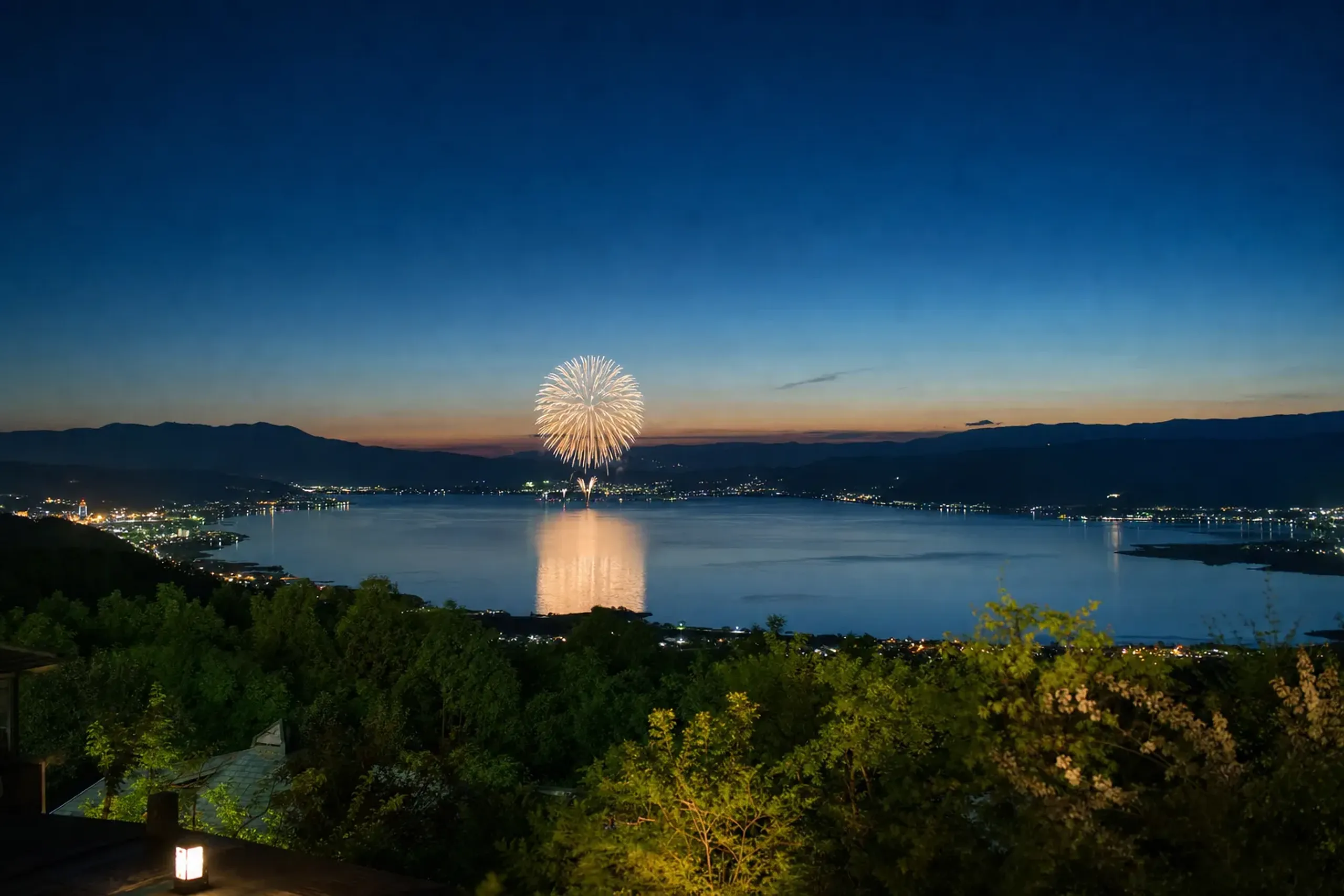 The mountain-ringed natural bowl of Lake Suwa at dusk, dark peaks encircling the 16-kilometer shoreline as a firework burst reflects off the surrounding slopes