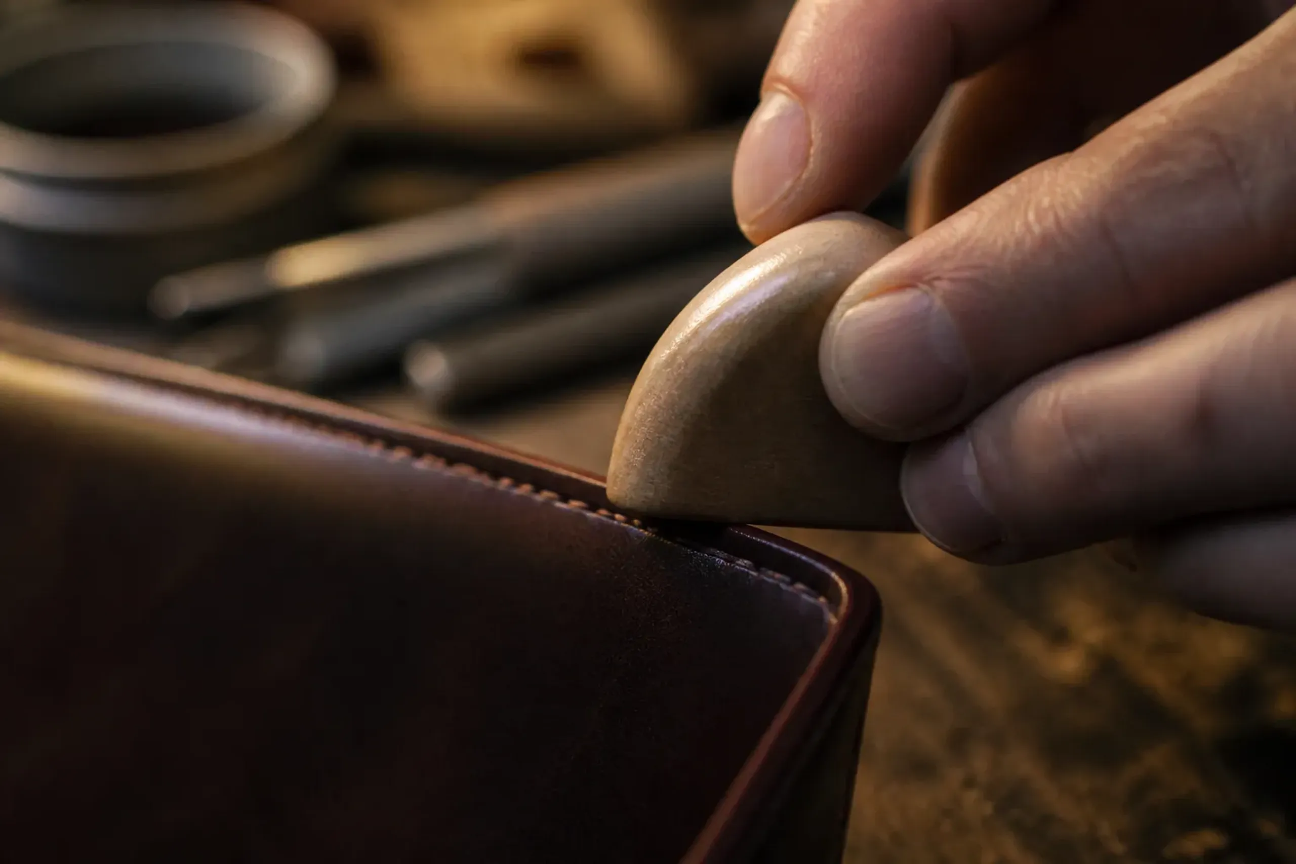 Hands burnishing the raw edge of a leather wallet with a wooden slicker tool