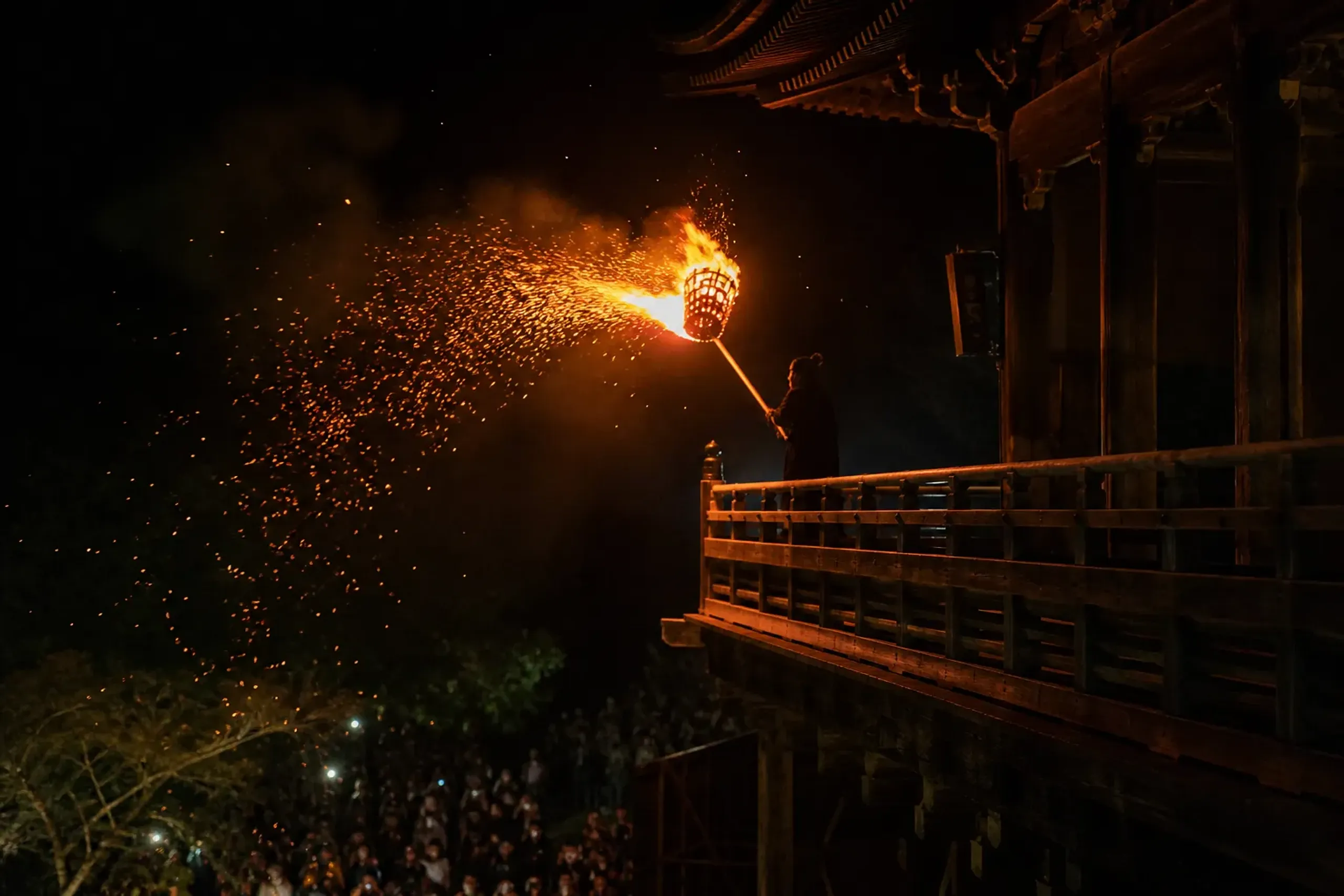 A dōji lay attendant carrying a burning six-metre cedar-leaf torch on a bamboo shaft along the wooden balcony of Nigatsu-dō, sparks scattering into the night over the hillside crowd below