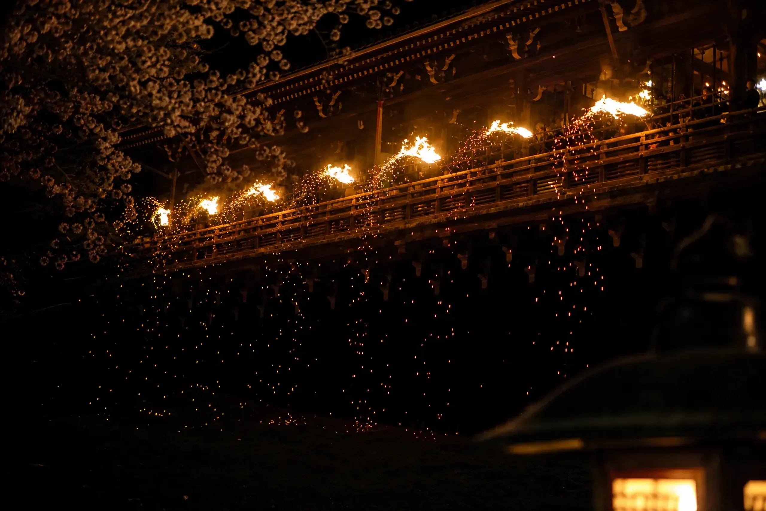 The illuminated balcony rail of Nigatsu-dō on an early-March weekday night, ten torches glowing along the old wooden threshold with open space on the lawn below