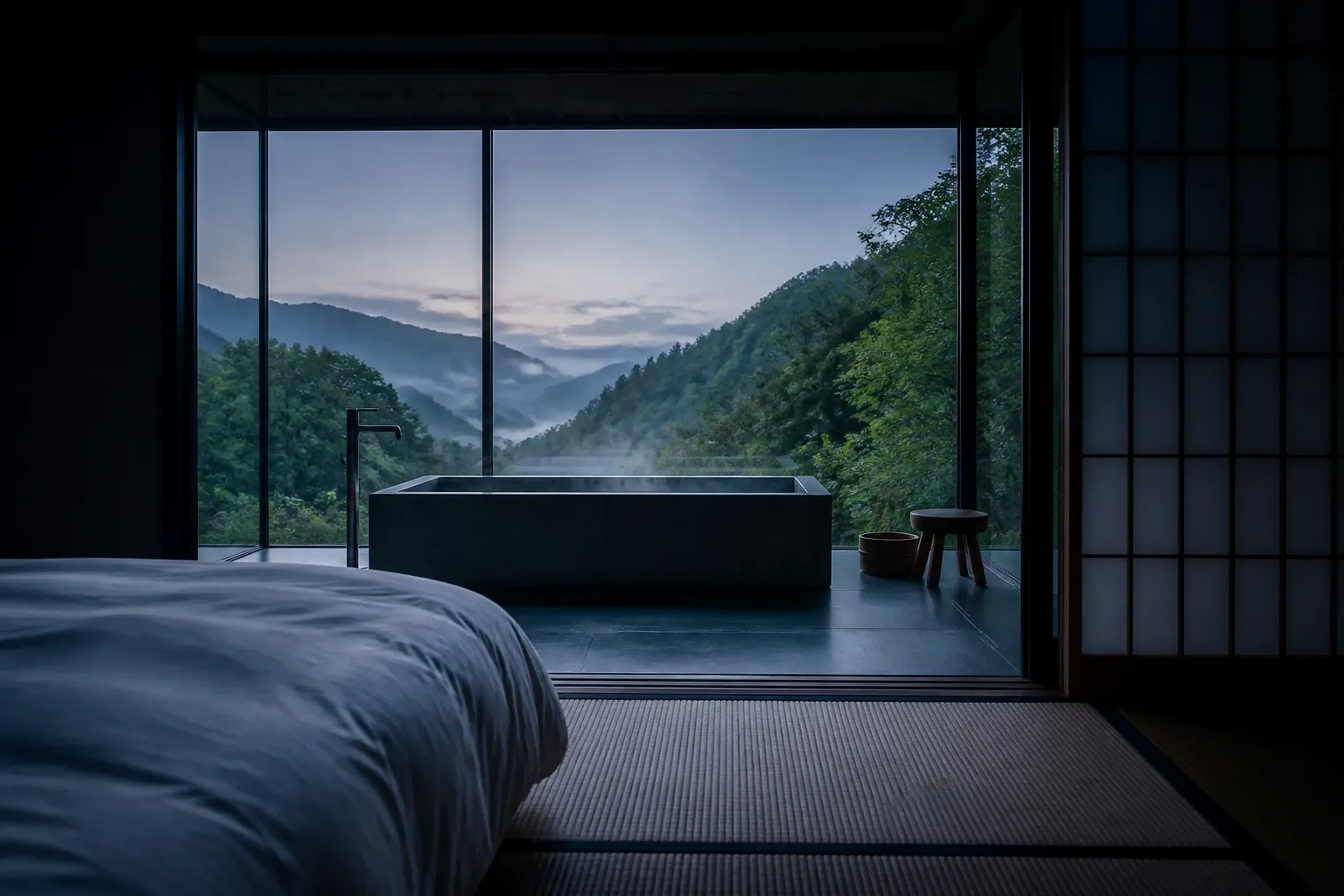A hybrid ryokan suite with tatami flooring, a low bed, and an in-room stone soaking tub facing a misty valley