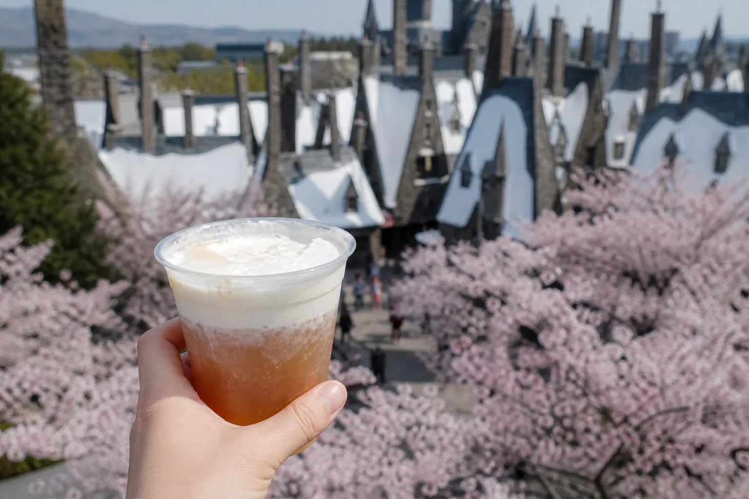 Hand holding a butterbeer-style drink with Hogsmeade lights blurred in the background at night