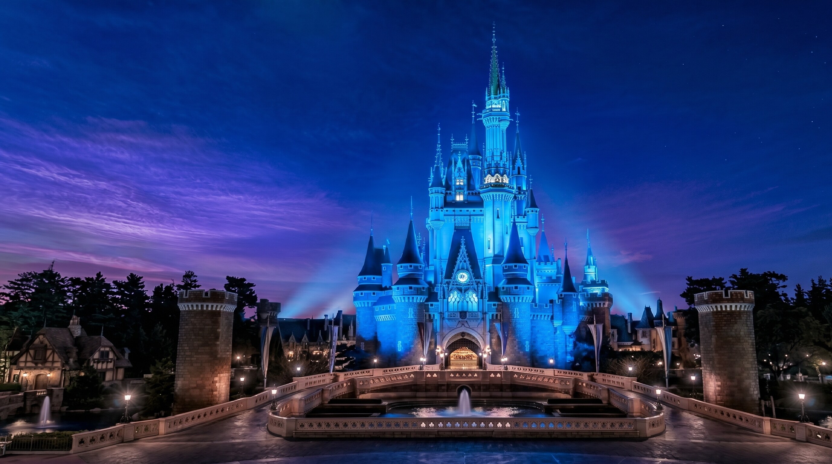 Disney castle illuminated in blue at night with a purple-tinged sky and surrounding trees in the foreground.