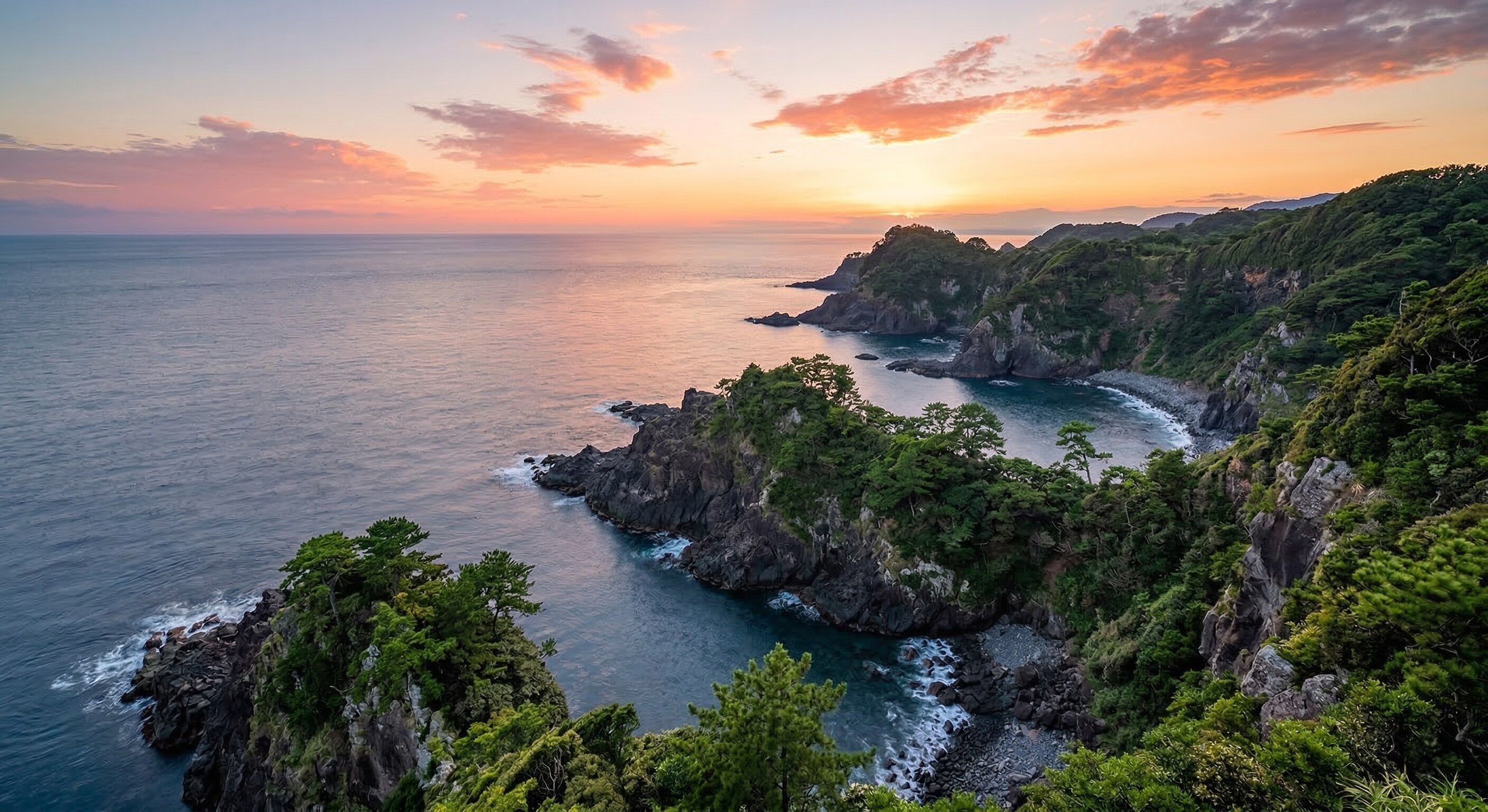 A dramatic view of the rugged Izu Peninsula coastline meeting the calm ocean waters during a vibrant sunset.