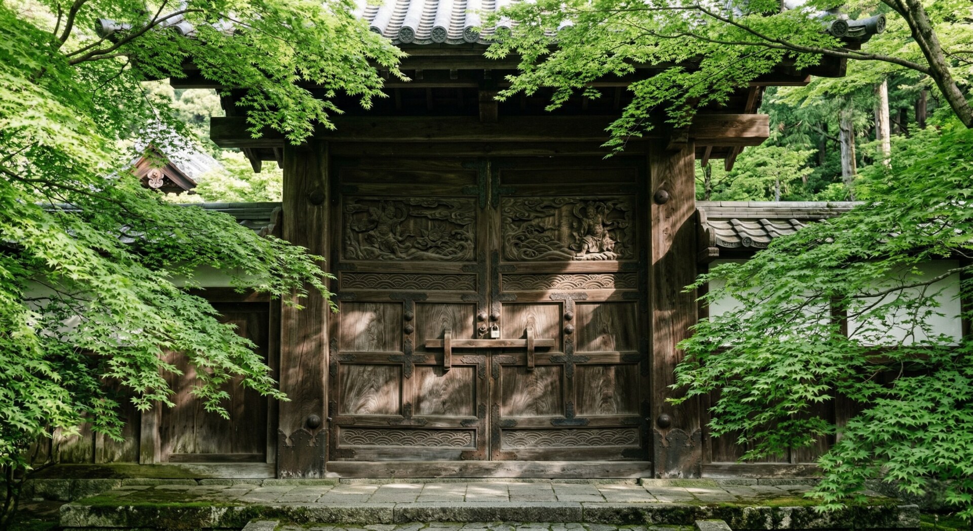Heavy, ancient wooden doors of a Kyoto temple closed to the public, surrounded by lush green maple trees.