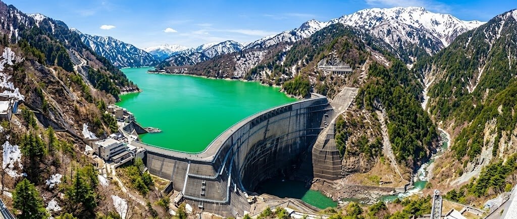 Panoramic view of a concrete dam creating a bright green reservoir in a valley surrounded by snow-capped mountains and pines.
