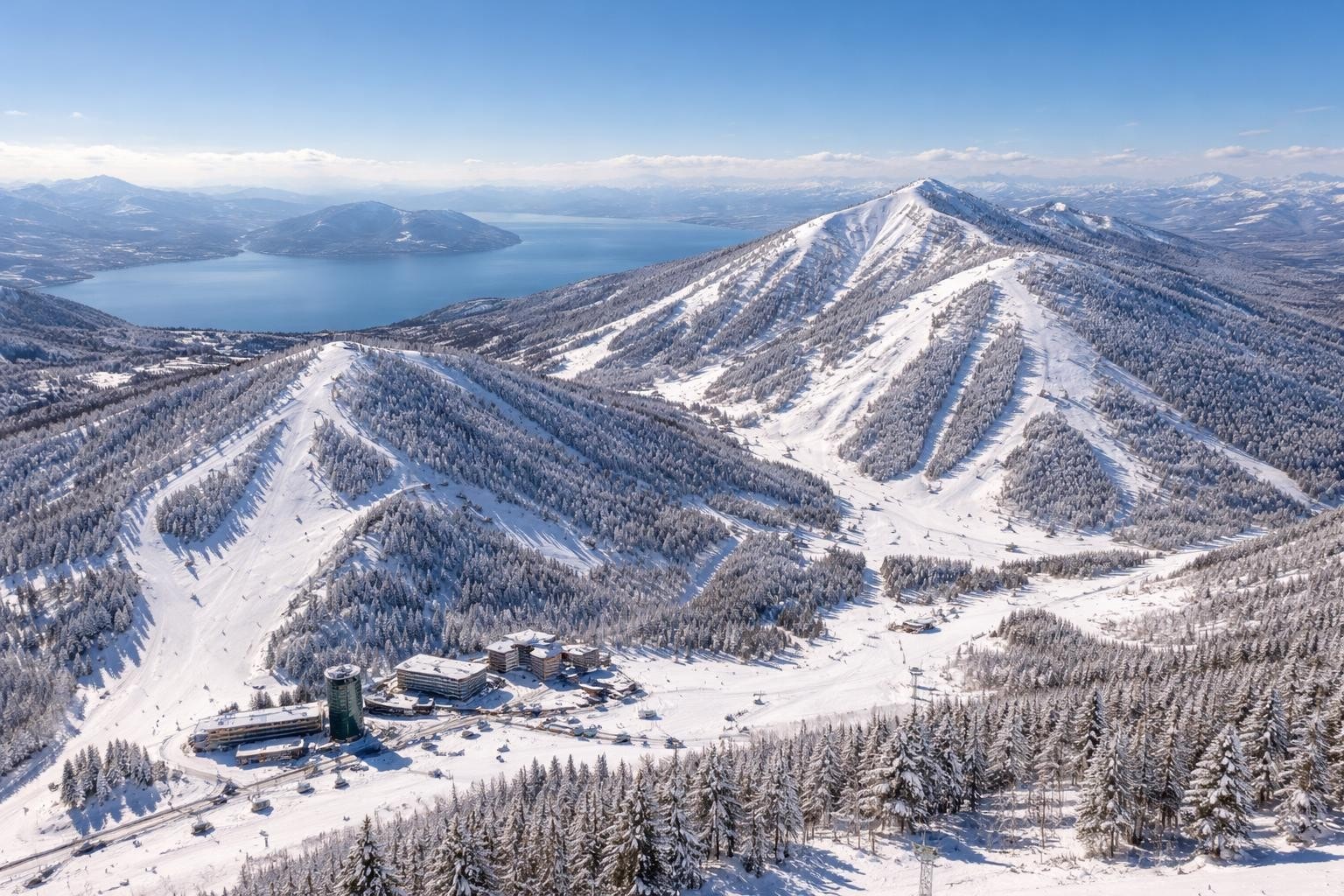Snow-covered alpine mountains with a blue lake in the distance and a ski resort complex at the base, under a clear sky.