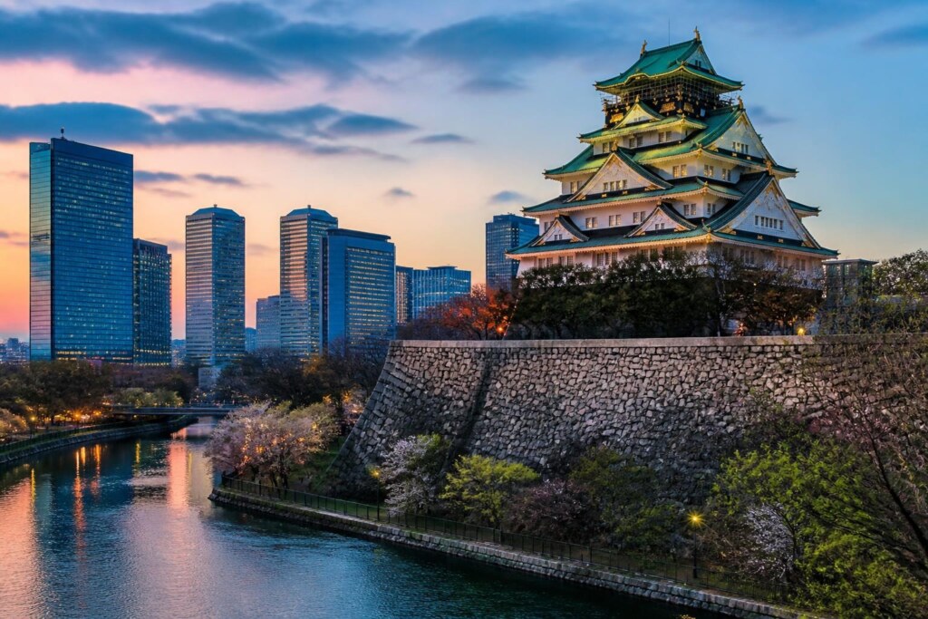 Osaka Castle atop a stone wall beside a river with a modern city skyline at sunset/sunrise behind it.