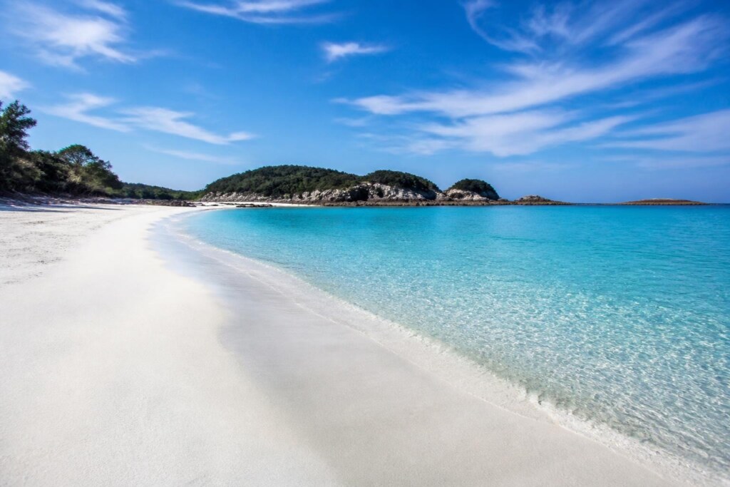 White sandy beach curves along turquoise water, with green, rocky islets in the distance under a blue sky. Readable shoreline with gentle waves.