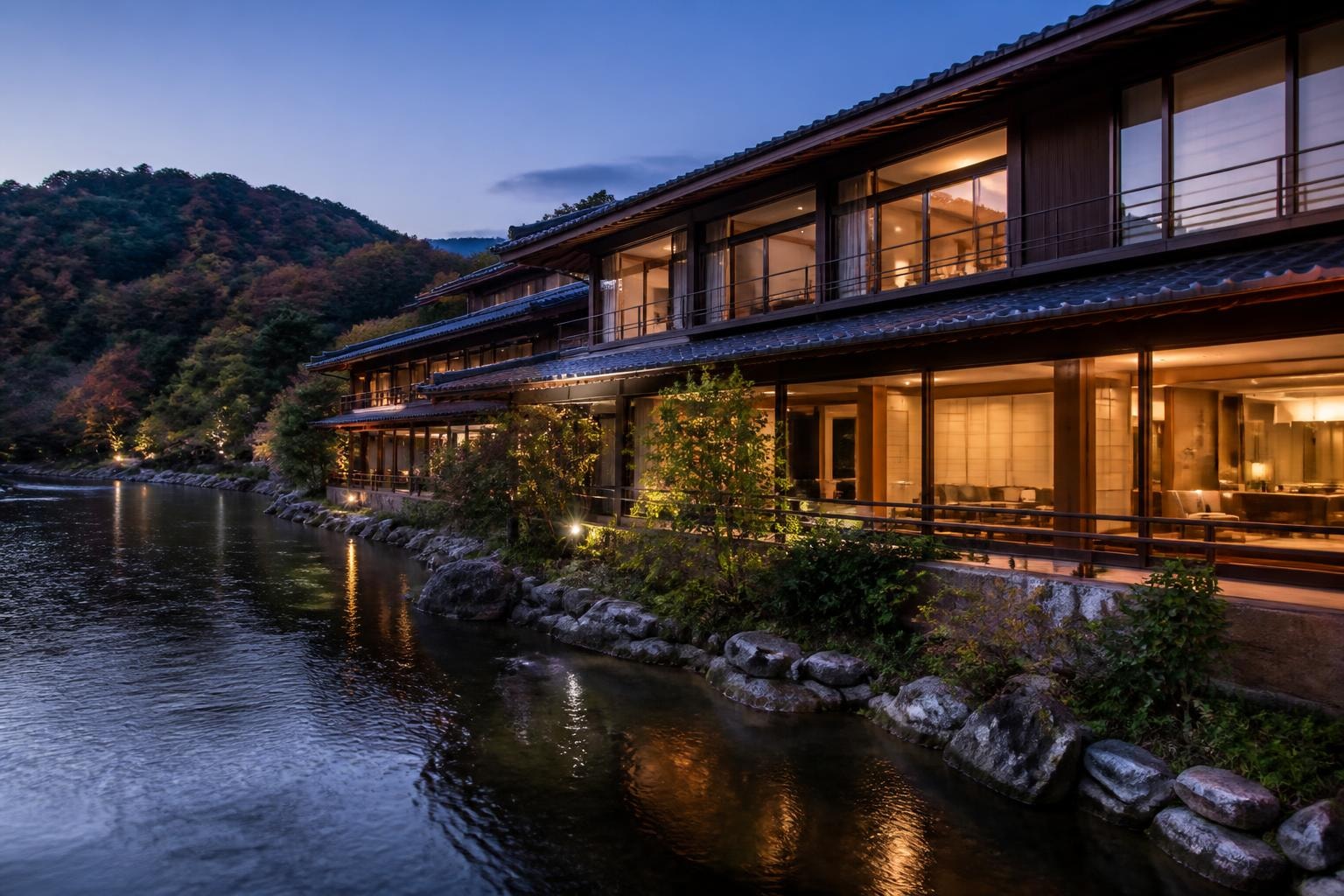 Evening view of a riverside hotel with warmly lit glass walls and wooden balconies along a rocky shoreline.