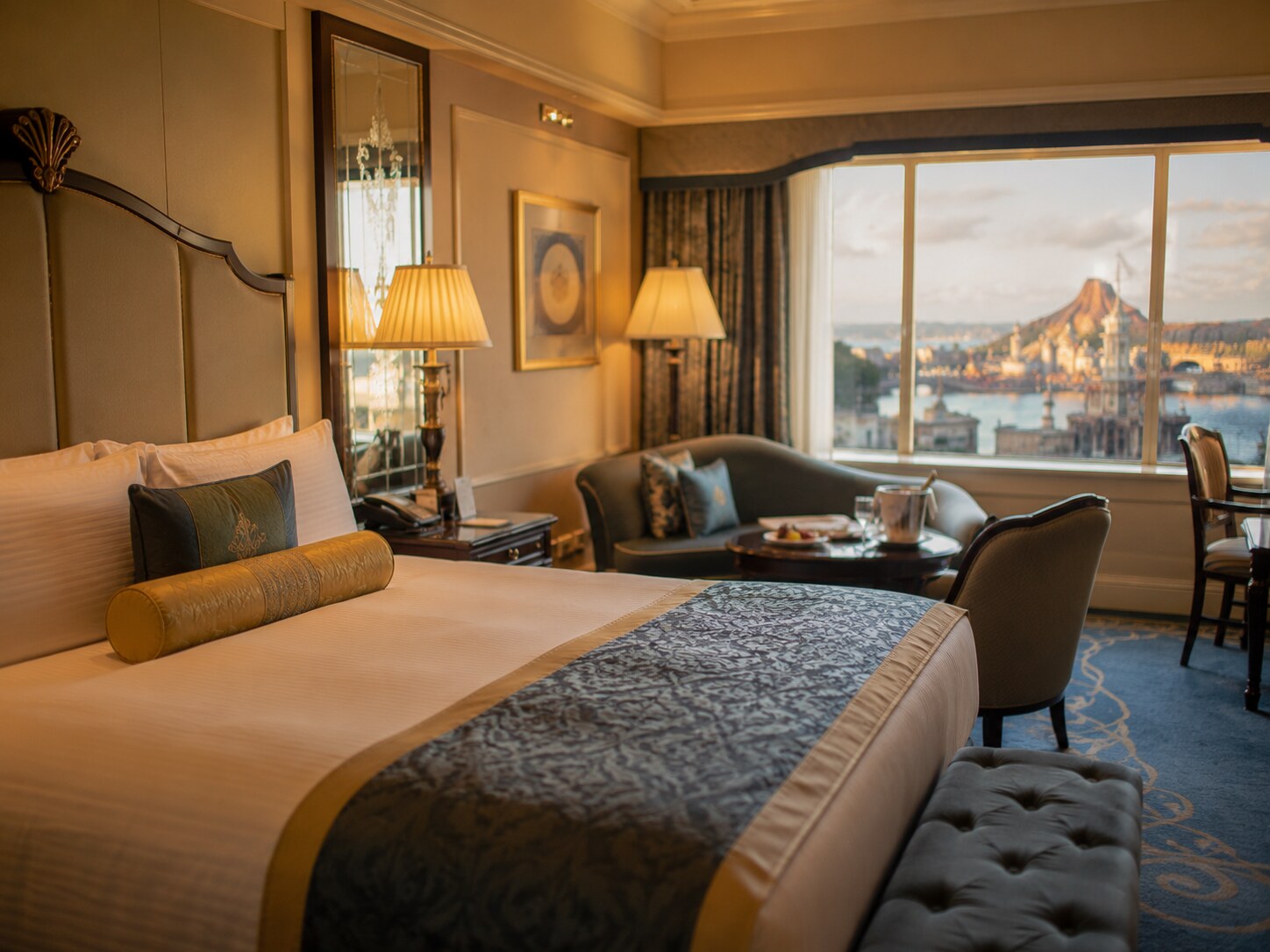 Luxury hotel room with a large bed, decorative pillows, and an ornate headboard; warm lamps illuminate a seating area by a window with a city and water view outside.