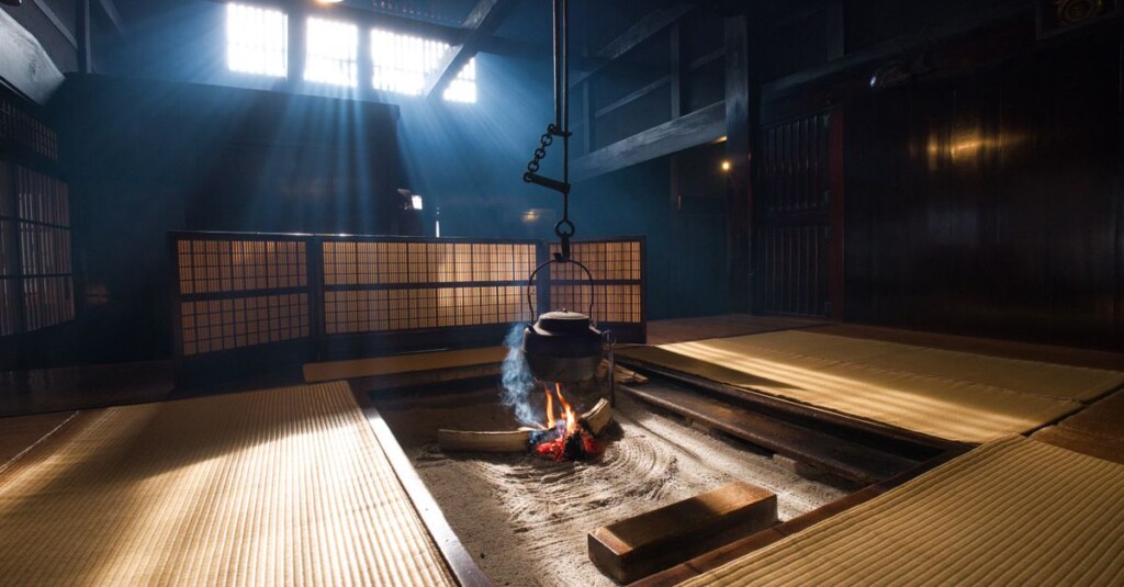 Traditional Japanese irori hearth with a hanging kettle over a wood fire, surrounded by tatami mats in a dim room.