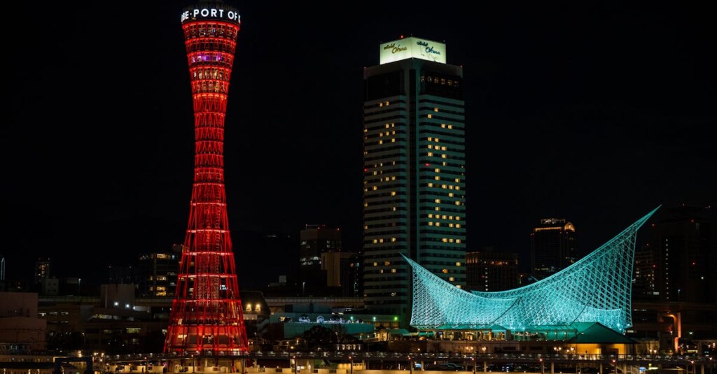 Night view of a city with a tall red lattice tower on the left and a blue illuminated sail-shaped structure on the right against a dark sky.