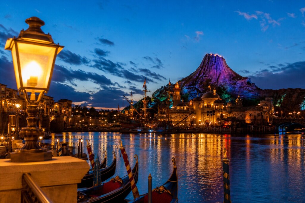 Evening harbor scene with gondolas, ornate lamps, and a colorful, lit volcano in the background.