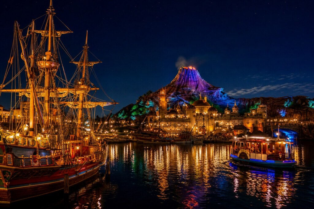 Night harbor with a tall-masted pirate ship on the left, a glowing castle and volcanic mountain center, and a lit sightseeing boat on the water, with colorful reflections.