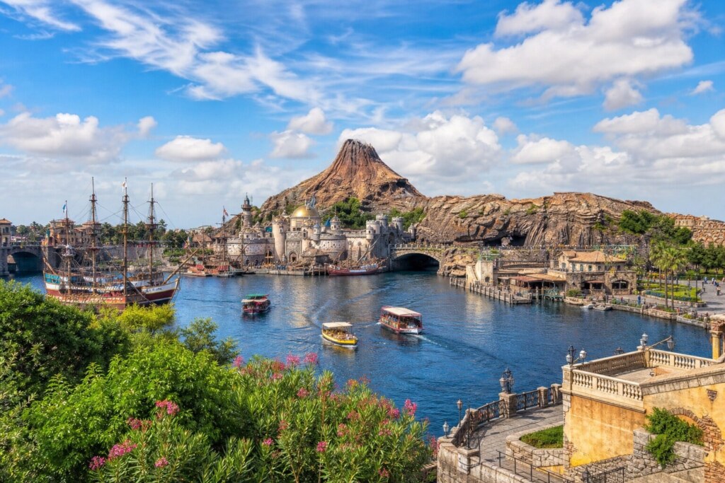 Waterfront scene in a theme park: colorful boats on a blue canal with a stone bridge and a castle-like harbor, and a rocky cone-shaped volcano in the background.