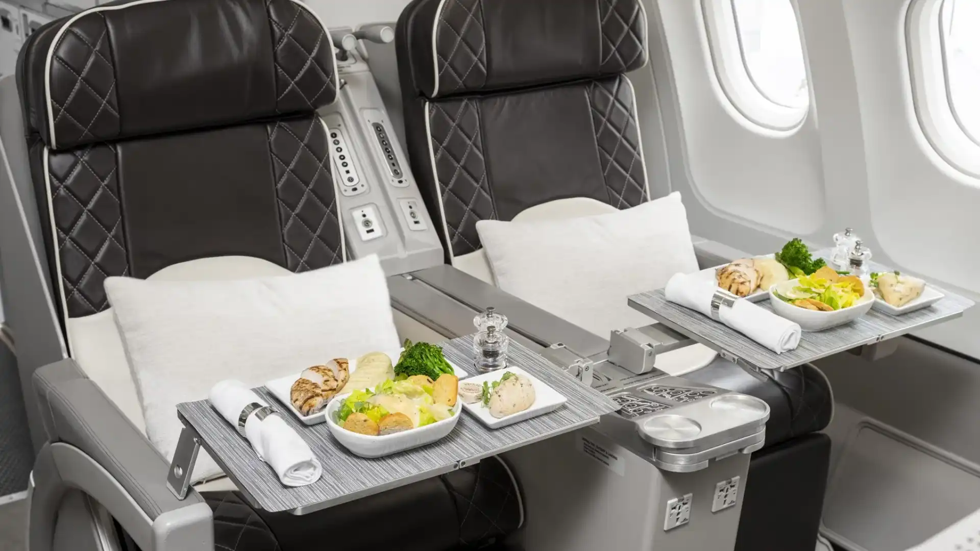 Close-up of two premium dark brown leather seats with diamond stitching next to a window in an Airbus A340-300. Deployed tray tables hold elegantly plated gourmet meals of chicken, salad, and vegetables, complete with silver napkin rings and salt and pepper shakers.