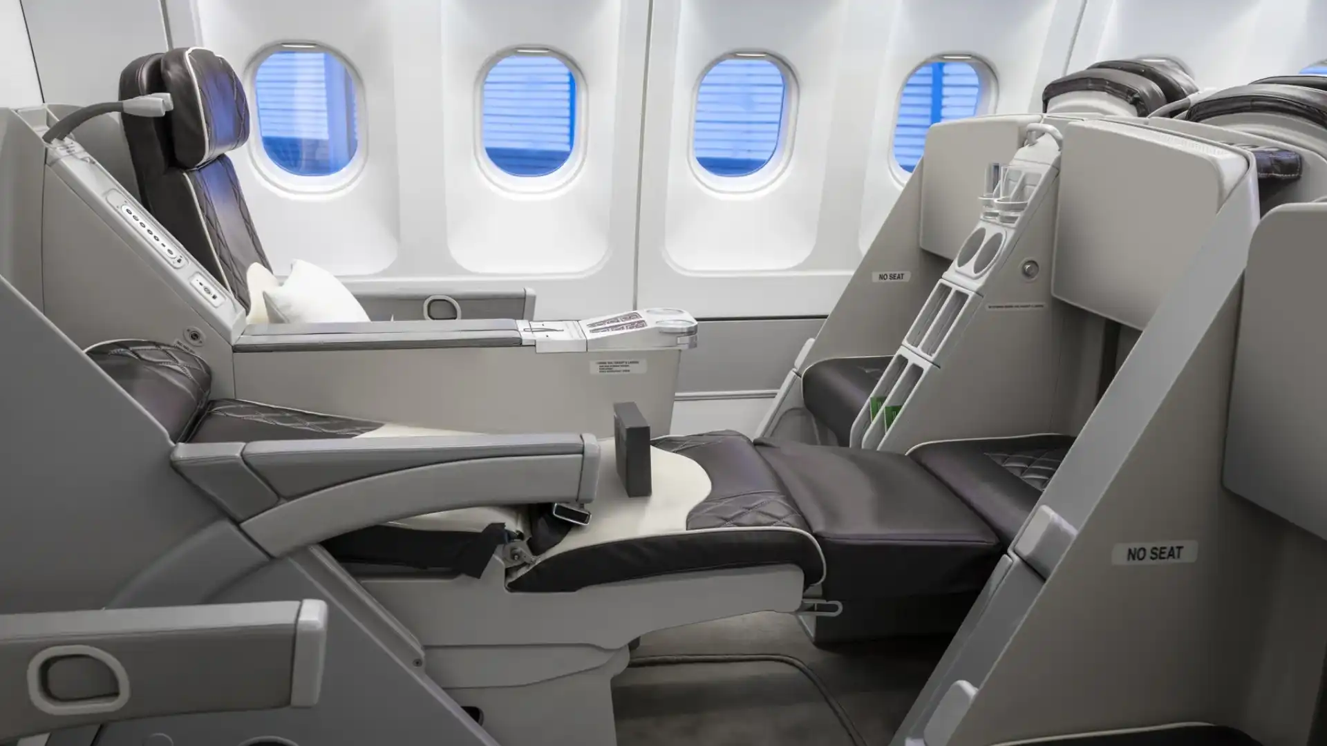Side view of two dark brown leather seats with diamond stitching in an Airbus A340-300. The closest seat is fully reclined into a flat bed position, connecting with a padded footrest. Four cabin windows with blue shades drawn down are visible in the background.
