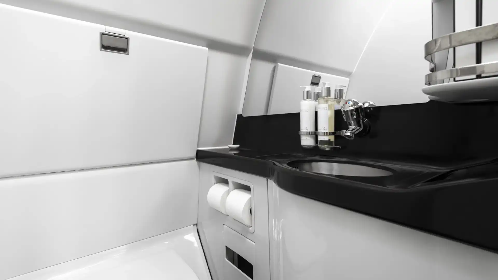 Interior view of an Airbus A340-300 VIP lavatory, featuring a modern design with clean white paneled walls, a glossy black countertop with an integrated stainless steel sink, a large mirror, and premium toiletries in a chrome dispenser.