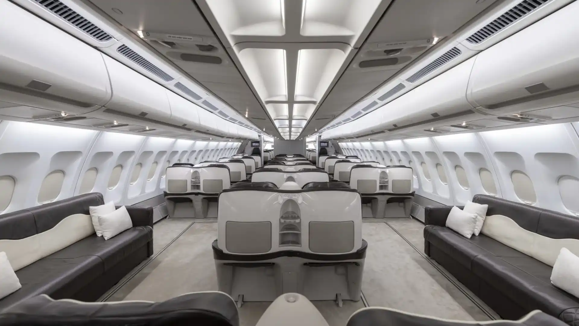 Interior view of a luxurious Airbus A340-300 VIP cabin, featuring long dark brown leather sofas with white pillows along the outer walls and premium, pod-style seats in the center aisle, illuminated by bright overhead lighting.