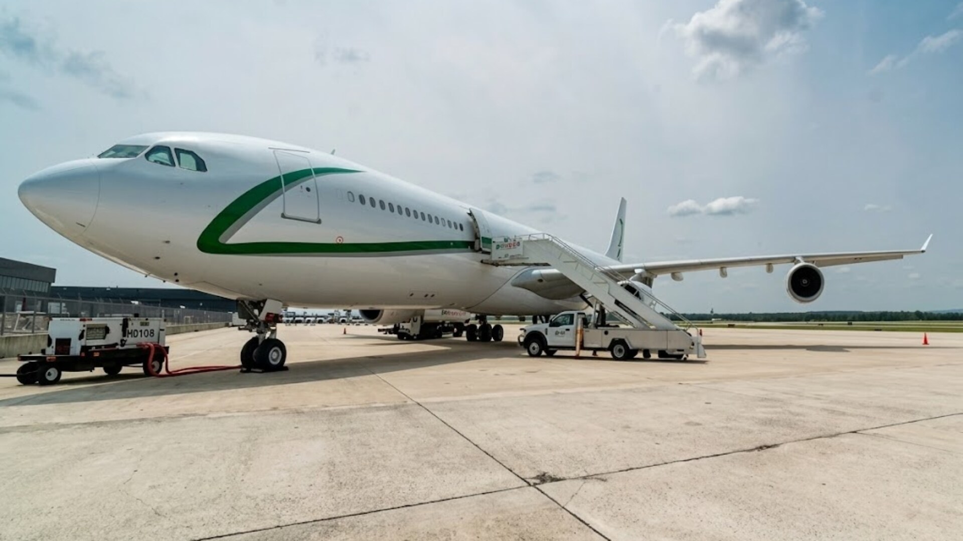 A wide-angle exterior view of a white Airbus A340-300 VIP aircraft parked on a concrete tarmac under a partly cloudy sky, featuring green accent stripes on the fuselage.